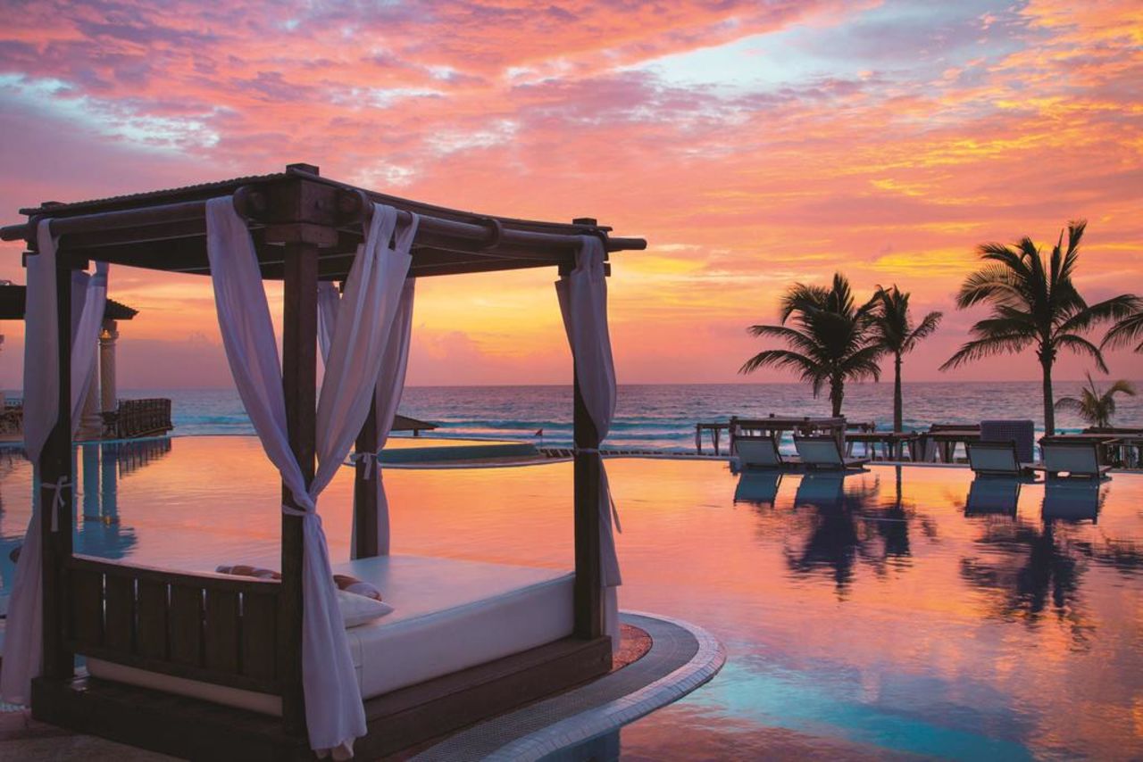 Large bed outside with a pool overlooking the ocean at sunset in cancun